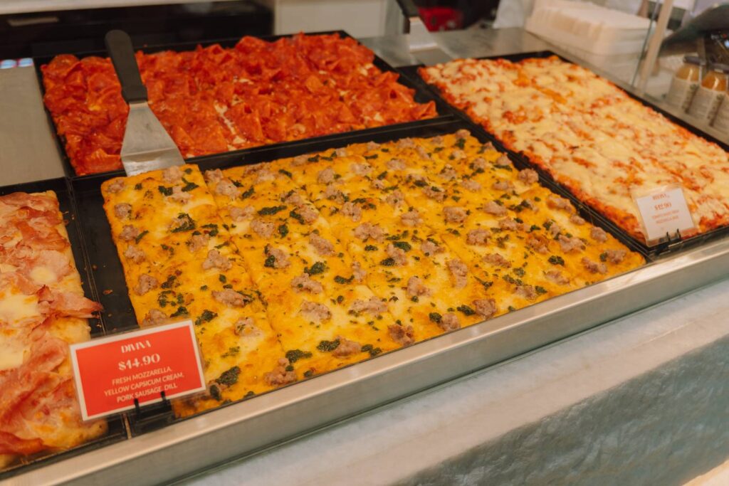 Eine Auswahl an verschiedenen frisch zubereiteten, rechteckigen Pizzastücken mit Belägen wie Paprikacreme, Wurst und Salami in der Auslage des New Sydney Fish Market.
