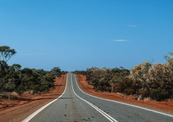 Eine schnurgerade Asphaltstraße führt durch die rote Erde und karge Vegetation des Outbacks unter einem strahlend blauen Himmel auf dem Roadtrip von Perth nach Exmouth.