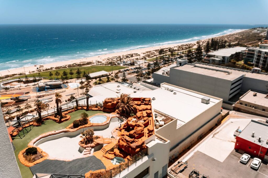 Eine erhöhte Aussicht auf die felsig gestaltete Dachterrasse mit Poolanlage des Rendezvous Hotels im Vordergrund, dahinter der weiße Sandstrand von Scarborough in Perth und der tiefblaue Indische Ozean unter strahlend blauem Himmel.