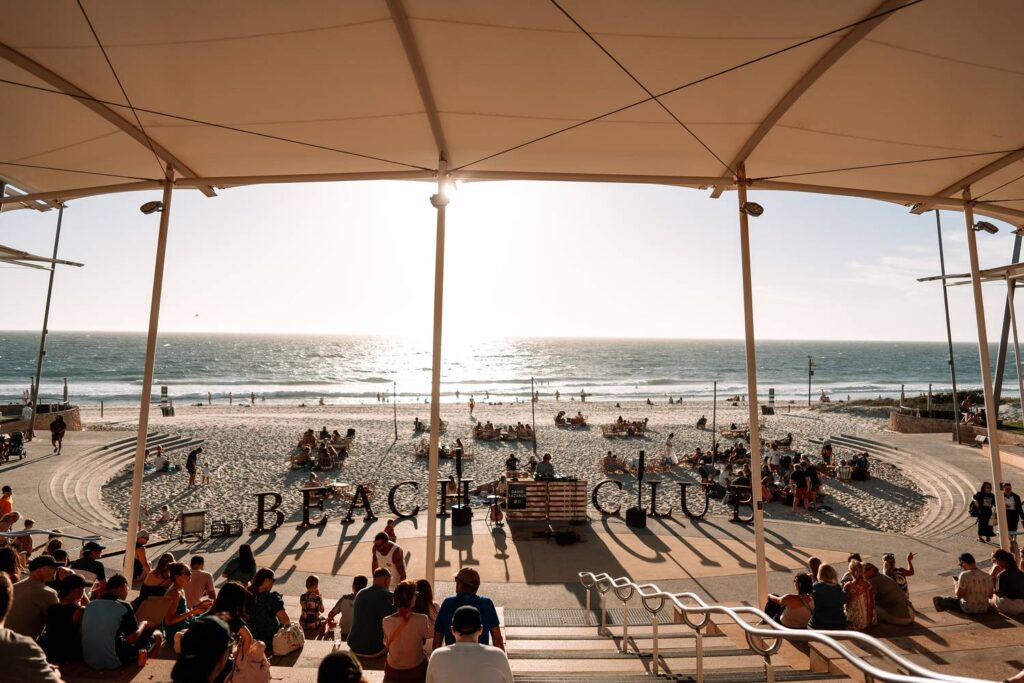 Unter einem großen weißen Sonnensegel sitzen zahlreiche Menschen im Amphitheater am Strand von Scarborough und genießen bei strahlendem Sonnenschein die entspannte Atmosphäre der Sunset Markets mit Blick auf das Meer.