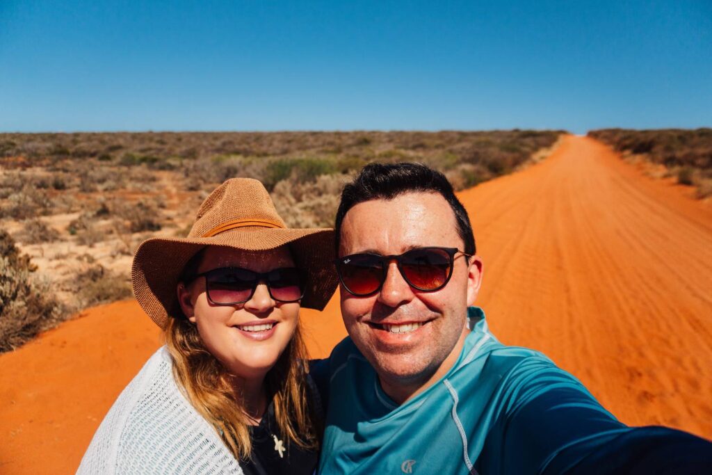 Daniela und Mario von Auszeit Abenteuer lächeln bei einem Selfie auf einer leuchtend roten Sandpiste im Francois-Peron-Nationalpark unter strahlend blauem Himmel.