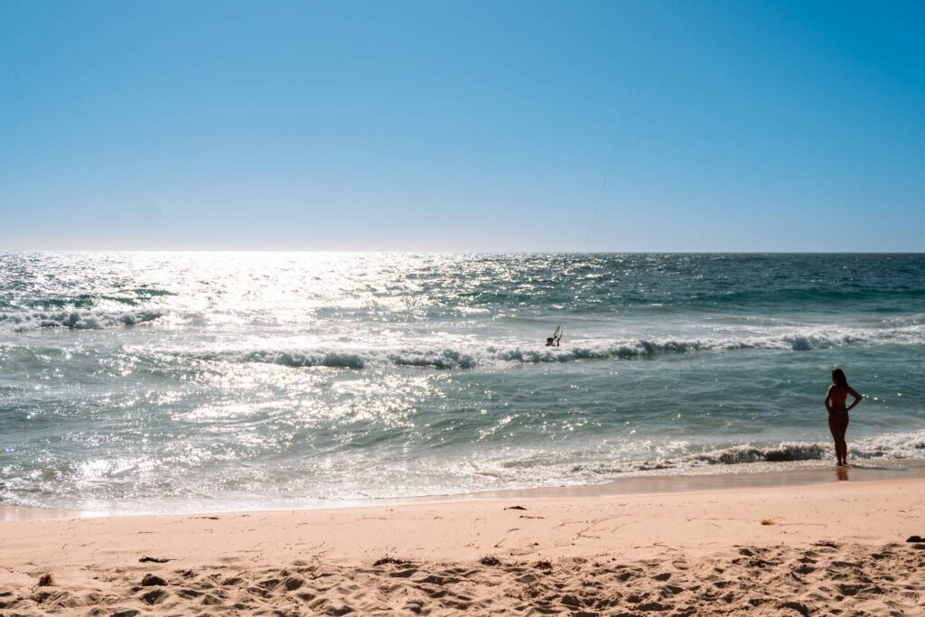 Eine Frau steht am sandigen Strand von Scarborough bei Perth und blickt auf das im hellen Sonnenlicht glitzernde Meer unter einem wolkenlosen blauen Himmel.
