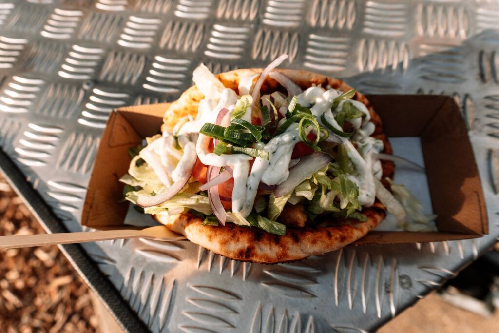 Ein frisch zubereitetes, reich belegtes Pita-Wrap mit knackigem Salat, roten Zwiebeln und weißer Knoblauchsauce, serviert in einer Pappschale bei den Sunset Markets in Scarborough im warmen Abendlicht.