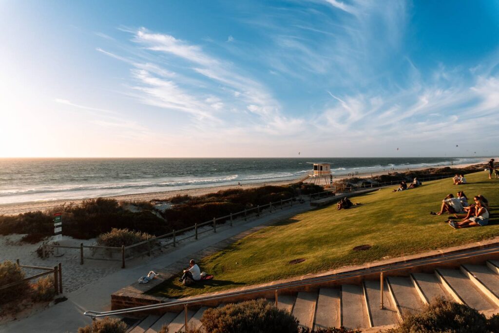 Besucher sitzen entspannt auf der grünen Wiese des Sunset Hill am Scarborough Beach in Perth und genießen die warme Abendsonne über dem Meer während der Sunset Markets.
