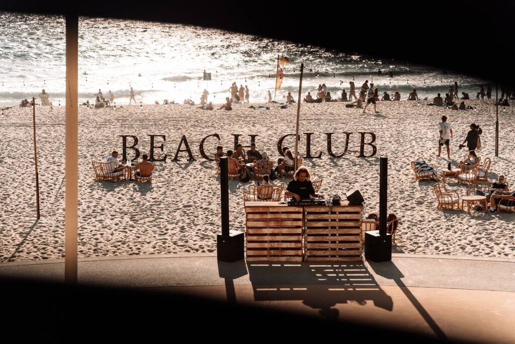 Ein DJ legt während des Sunset Markets am Strand von Scarborough in Perth an einem Mischpult aus Holzpaletten auf, im Hintergrund steht der große Schriftzug BEACH CLUB vor zahlreichen Besuchern und dem glitzernden Meer im Gegenlicht.