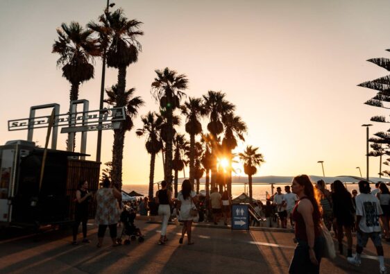 Zahlreiche Menschen flanieren im warmen Gegenlicht des goldenen Sonnenuntergangs an den Palmen der Scarborough Sunset Markets in Perth vorbei.