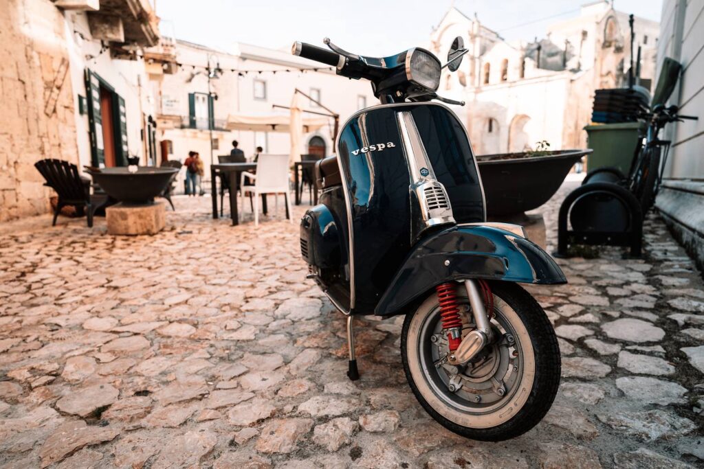 Eine dunkelblaue Vintage-Vespa parkt auf einem sonnigen Kopfsteinpflasterplatz in der historischen Altstadt Sassi von Matera.