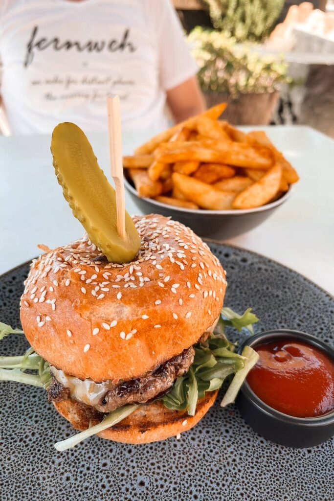 Ein appetitlicher Hamburger mit einer großen Gewürzgurke auf einem Holzspieß, serviert auf einem dunklen Teller neben Ketchup und einer Schale goldgelber Pommes in Jervis Bay.