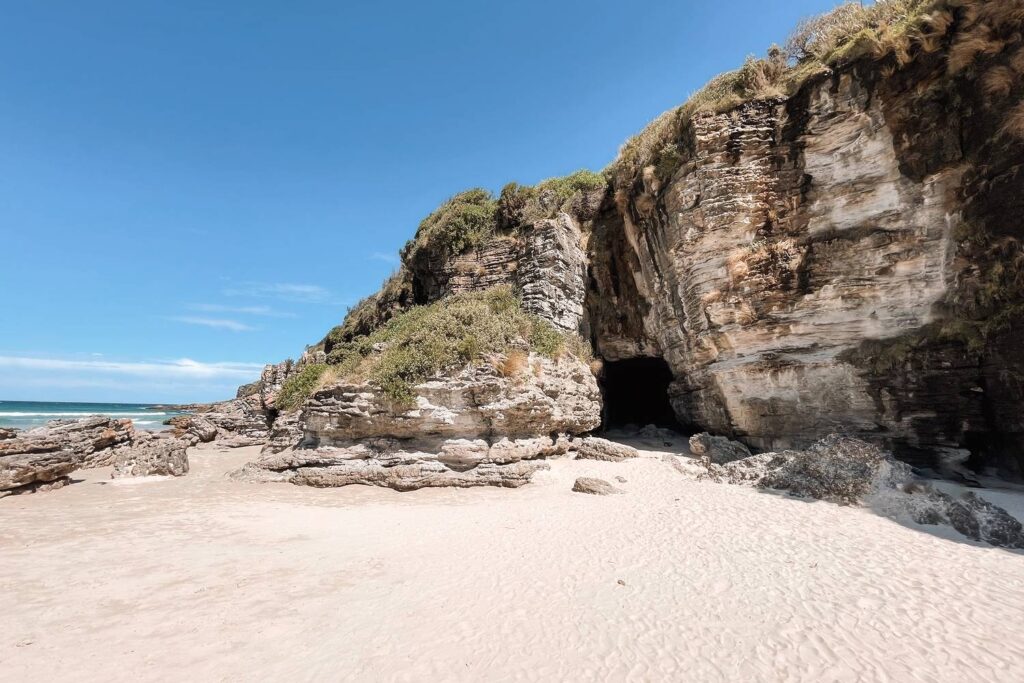 Eine markante, dunkle Felshöhle in den hellen Sandsteinklippen direkt am feinsandigen Cave Beach in Jervis Bay unter einem klaren, blauen Himmel.