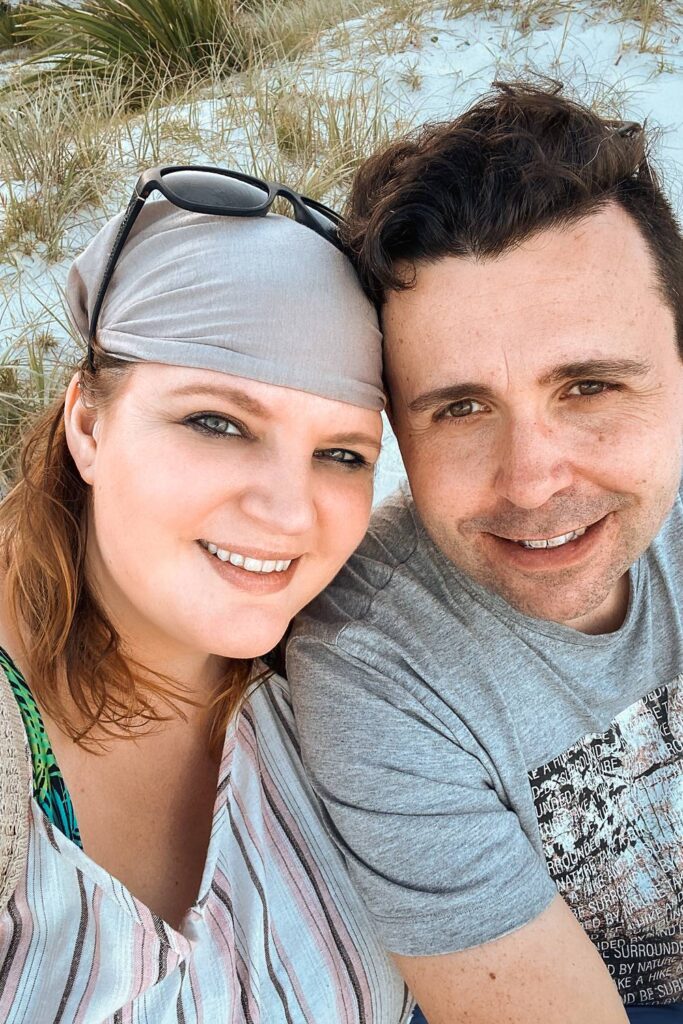 Daniela und Mario lächeln glücklich für ein Selfie am feinen, weißen Sandstrand von Jervis Bay vor einer Kulisse aus grünem Dünengras.