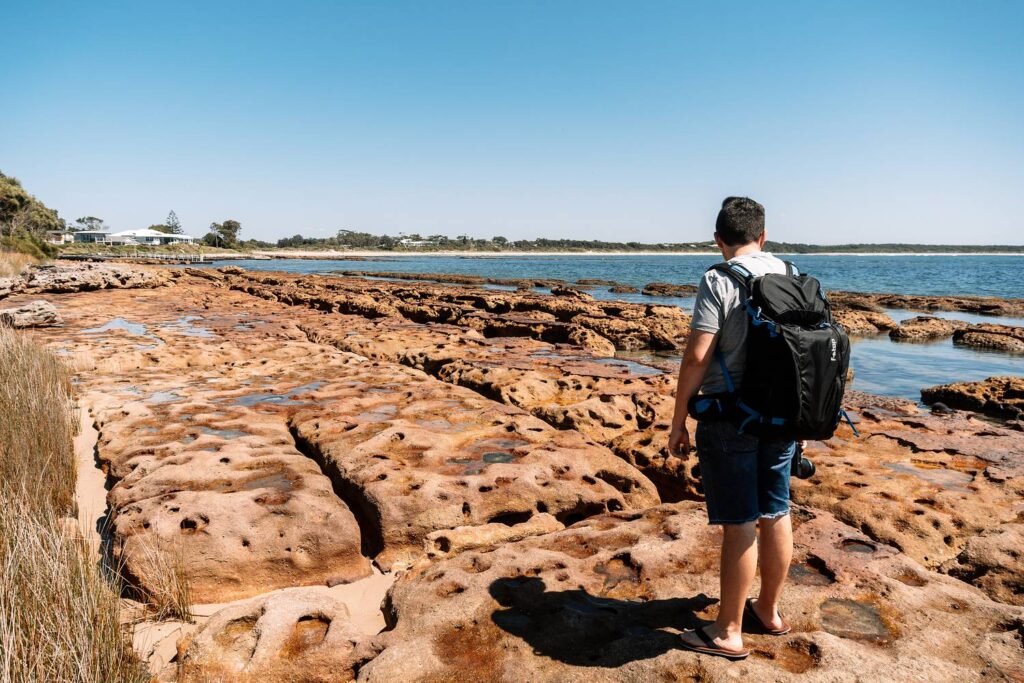 Mario steht mit einem großen Kamera-Rucksack auf den markanten, rötlichen Felsformationen an der Küste von Jervis Bay und blickt an einem sonnigen Tag auf das blaue Meer.