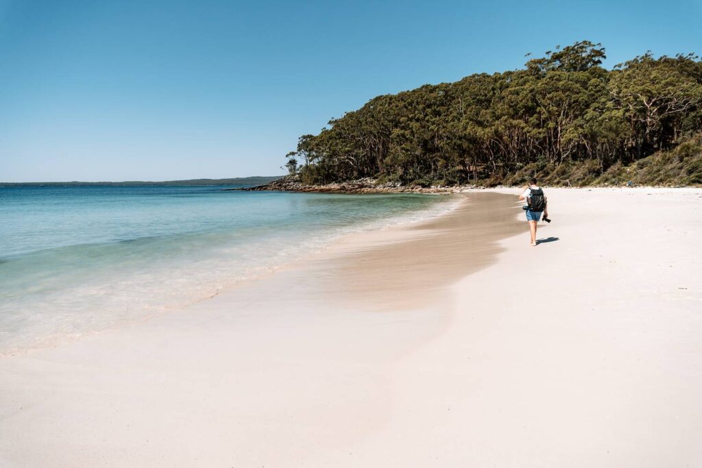 Mario läuft mit einem Rucksack und einer Kamera entlang des schneeweißen Sandstrandes an der Jervis Bay in Australien, gesäumt von kristallklarem, türkisfarbenem Wasser und einem dichten grünen Wald unter strahlend blauem Himmel.