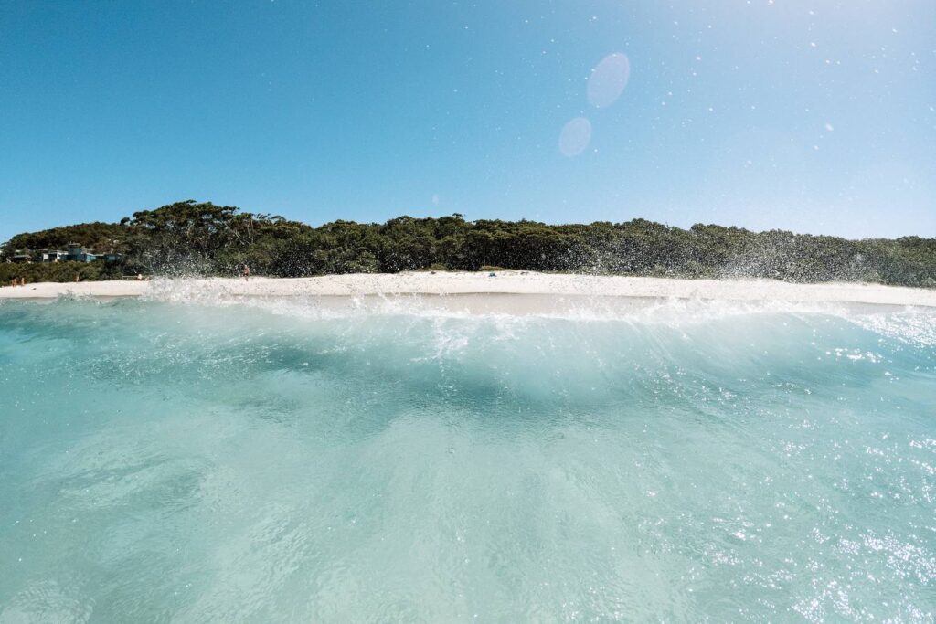 Eine kleine, sich brechende türkisfarbene Welle mit glitzernder Gischt im kristallklaren Wasser vor dem weißen Sandstrand von Jervis Bay unter strahlend blauem Himmel.