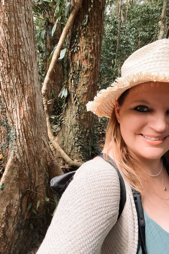 Daniela lächelt mit einem Strohhut während einer Wanderung vor den dichten, urigen Baumstämmen des Daintree-Regenwaldes in Queensland.