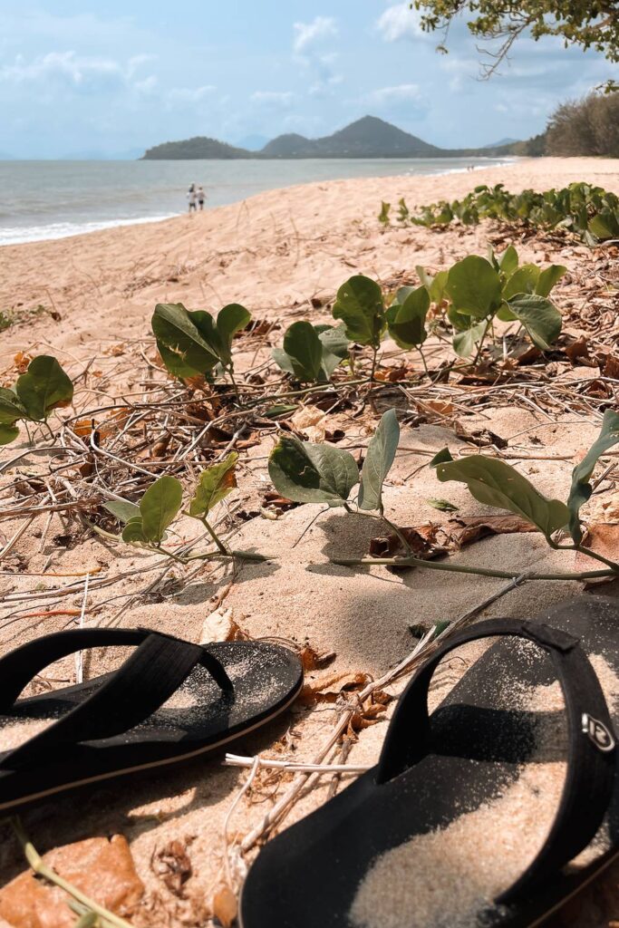 Schwarze Flip-Flops liegen im feinen Sand eines sonnigen australischen Strandes mit Blick auf das blaue Meer und grün bewachsene Hügel im Hintergrund.