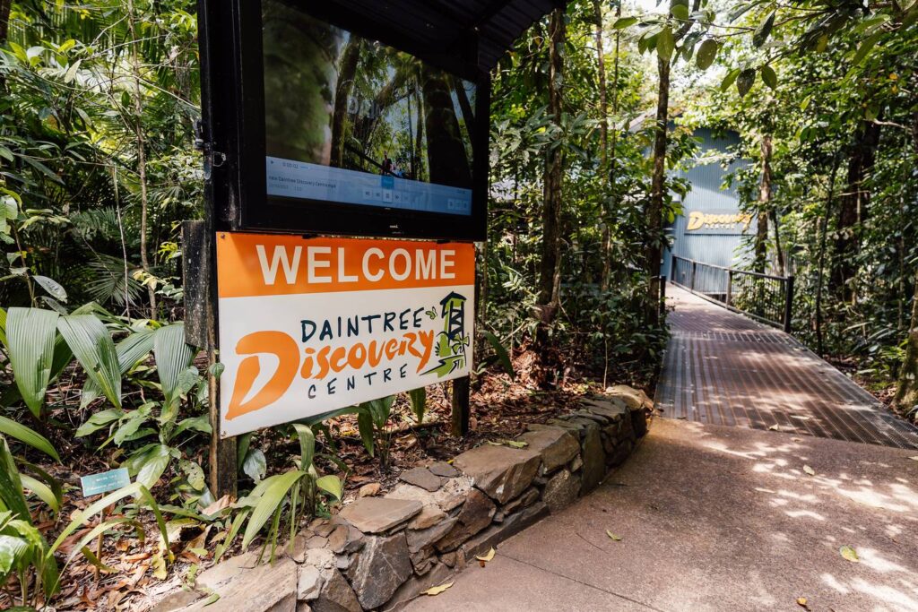 Ein Willkommensschild mit Informationsbildschirm am Eingang zum Daintree Discovery Centre, von dem aus ein Holzsteg in den dichten tropischen Regenwald führt.