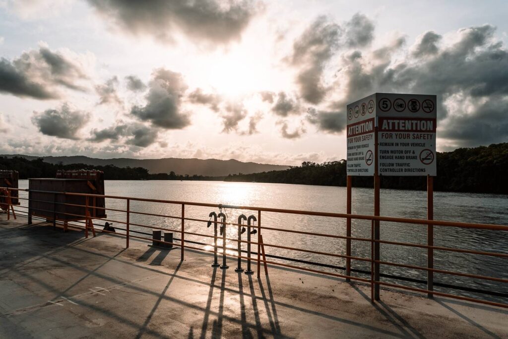 Blick vom Deck der Daintree Ferry mit Sicherheitsschildern über das sonnenbeschienene Wasser des Flusses auf den dichten Regenwald im atmosphärischen Gegenlicht.