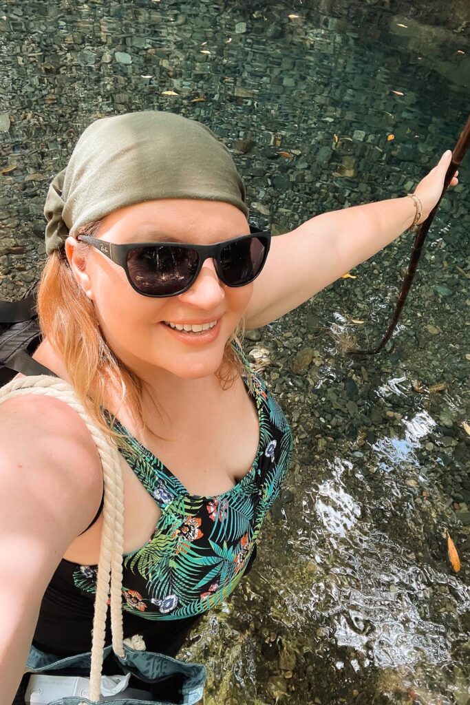 Daniela lächelt mit Sonnenbrille und grünem Kopftuch bei einer Erfrischung im glasklaren Wasser eines Regenwald-Creeks an der Heritage Lodge in Daintree.