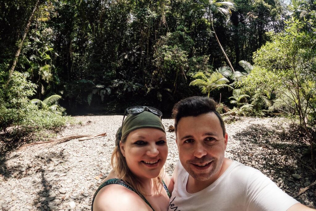 Daniela und Mario lächeln für ein Selfie am sonnigen, steinigen Ufer eines Regenwald-Creeks nahe der Heritage Lodge im dichten Daintree-Nationalpark.