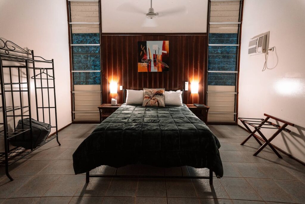 Gemütlich eingerichtetes Schlafzimmer der Heritage Lodge im Daintree-Regenwald mit bezogenem Doppelbett, warmer Wandbeleuchtung und großen Lamellenfenstern mit Blick ins Grüne.